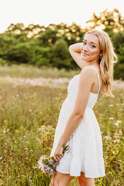 beautiful teen girl in a white dress holding a bouquet of flowers and playing with her hair captured by Ashley Kalbus Photography