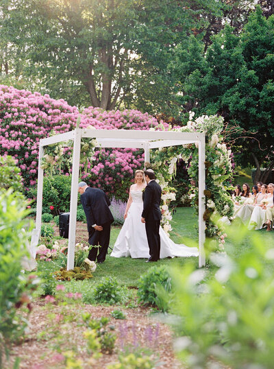 Glen Manor House | A couple stands under a floral-decorated white chuppah in a lush garden with pink flowers, during a wedding ceremony. Guests are seated in the background.