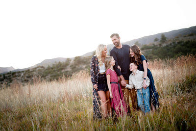 A family of 5 hold hands and laugh together in a field of tall grass