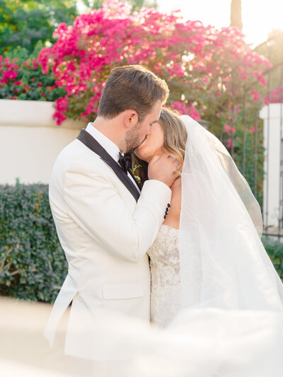 An elegant bride and groom sharing a romantic kiss amid vibrant bougainvillea at their destination wedding in Palm Springs, captured in a refined fine art style by Elizabeth Kane Photography—serving Philadelphia, New York, and the East Coast.