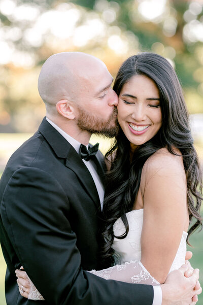 Close up photo of a groom kissing his bride on her cheek as she is smiling.