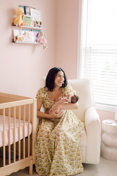 A mother smiles while holding her newborn baby in a cozy pink nursery, captured by a Northern Virginia newborn and family photographer during an in-home session.