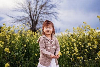 a huge field of wildflowers (mustard flowers) surrounds a new mom in a salmon maternity dress