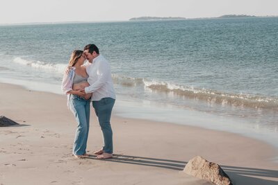Husband and wife posing on beach for maternity photoshoot.