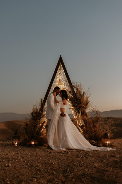 bride and groom stand in front of their wedding arch in the desert