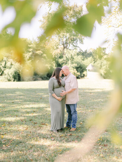 Expecting Mom and her husband snuggle during a maternity photo session surrounded by greenery at Southern Maryland farm.