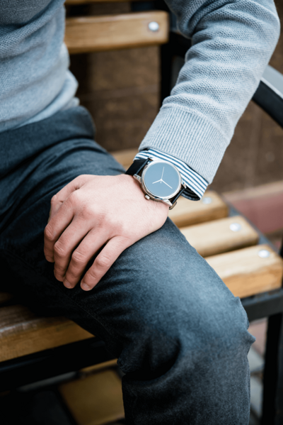 Close-up shot of a modern minimalist watch with a sleek black face and silver case, worn with a light gray sweater featuring striped cuff details and dark denim jeans. The lifestyle photography captures a casual yet sophisticated men's fashion accessory against a wooden bench background, demonstrating contemporary urban style.