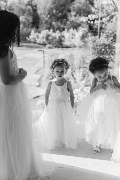 New England Wedding Photographer Pricing | Kelsey Sheehan Photography Timeless Rhode Island Weddings | Three children in white dresses stand together inside, with soft light filtering through a window. They appear joyful and curious.