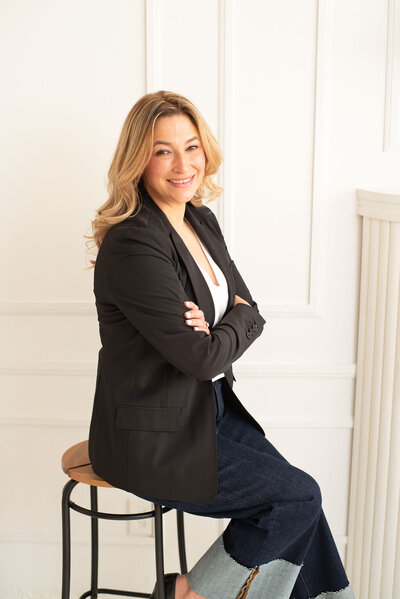 Laura Gatsos Young seated on a stool wearing a dark blazer and blue jeans