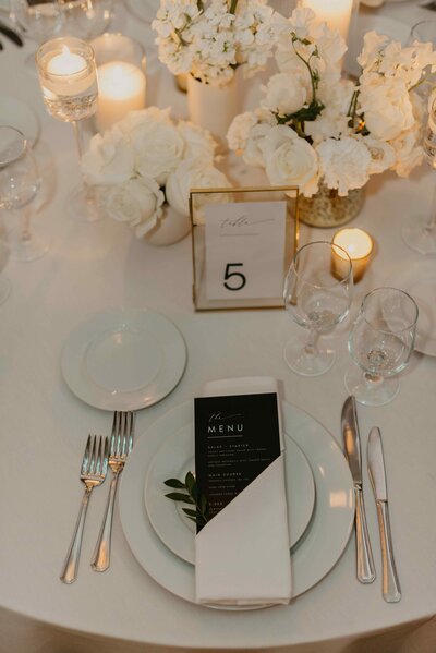 Elegant wedding table setting with white floral arrangements, candles, and a black and white menu card on a neatly folded napkin.