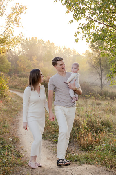 a family wearing whites and neutrals hold hands as they smile at each other and hold their 6 month old baby. The family walks through a grassy field with a foggy background 