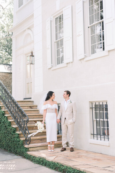 Bride and Groom holding hands in front of a historic building in downtown Charleston during their fashion forward elopement. Bride in a lace corset midi dress