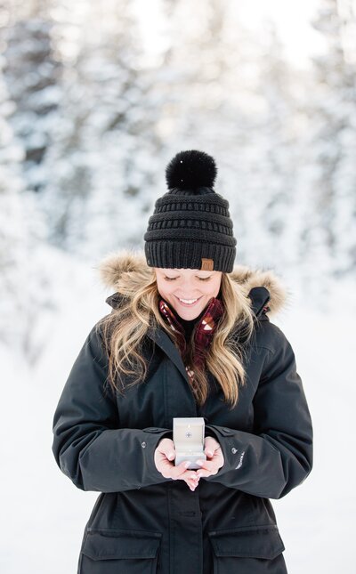 A woman in a black jacket and beanie smiles down at her engagement ring in the box in her hands