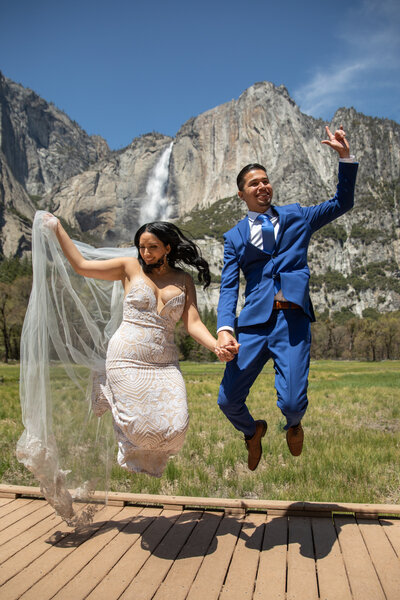 A newlywed couple celebrates their Yosemite elopement in front of Yosemite Falls, jumping with joy on a wooden boardwalk. Captured by Yosemite elopement photographers, this moment reflects the excitement of an adventurous wedding day.