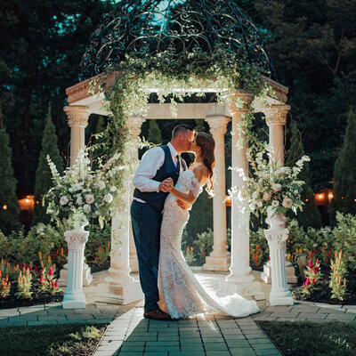 Bride and groom kissing under gazebo with ivy, flowers, and twinkle lights | Summer Night Wedding at The Hamilton Manor | Hamilton Township, New Jersey