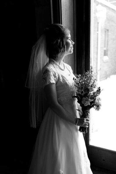A smiling bride holding her bouquet looks out a window as the light hits the front of her body.