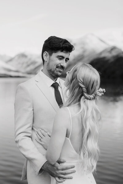 Groom looking lovingly at his bride who has long flowing hair. He is wearing a pale coloured suit and they are in front of a picturesque lake.