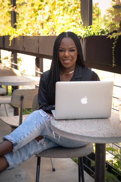 Rianne wearing a black top and jeans sitting a a table outside near a coffee bar. On the table in front her her is a silver macbook that's opened while she is smiling at the camera.