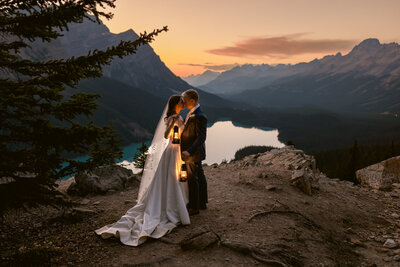 Bride and groom holding lanterns at Peyto Lake in Banff National Park during a warm summer sunset standing close together with a soft golden glow in the background captured as a romantic evening wedding portrait