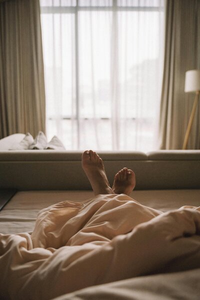 Cozy photo of person lying under a blanket on a bed with feet peeking out, facing a softly lit window.