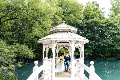 A full-length portrait of the couple in front of the white farmhouse, standing close together with foreheads touching. The bride’s veil drapes along the walkway in front of them, and both hold the pink and white bouquet between them, framed by lanterns and trimmed hedges.