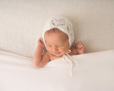 A beautiful baby girl in a white bonnet sleeps under a white blanket
