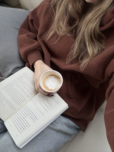 A woman holding a drink while reading a book. Showing that with anxiety therapy in Manhattan you can calm the inner chaos. An anxiety therapist can help you learn to relax and enjoy every day.