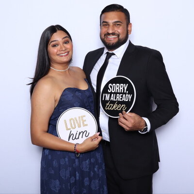 A smiling couple holding circular signs that read “Love him” and “Sorry, I’m already taken” in front of a white photo booth backdrop.