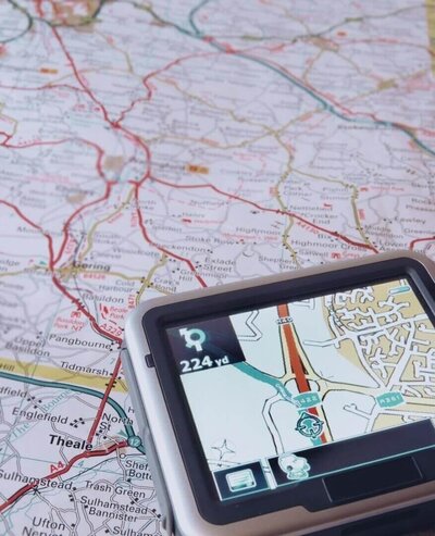 A close-up of a paper road map with a GPS device displaying a navigation route in the foreground, symbolising direction, planning, and finding your way.