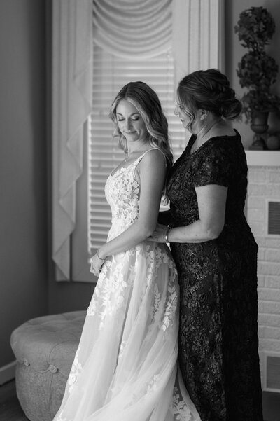 A bride and her mom getting dressed for the wedding. The mom is helping her daughter into her gown and whispering how proud she is. The bride is wiping away tears. 