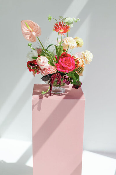 Pink and white floral arrangement with greenery sitting on a pink block in front of a white background
