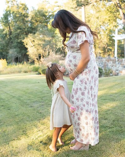 Pregnant mother in a floral dress shares a sweet moment with her young daughter in a sunlit field. A maternity session by Moments in Light Photography.