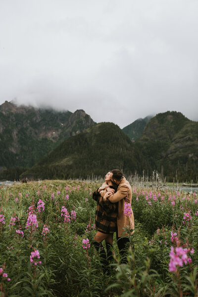 couple standing in a field of green and pink fireweed in front of stunning mountains during their alaska engagement photos in seward alaska