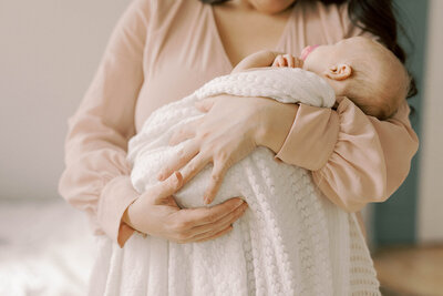 Moms arms holding newborn baby wrapped his white knitted blanket.