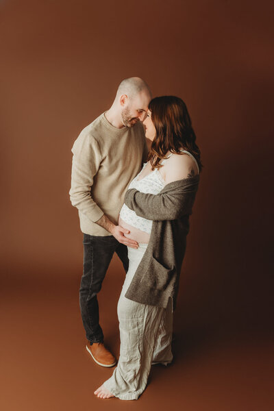 A couple stands close together, sharing an intimate moment. The woman, visibly pregnant, touches her belly, while the man gently leans in. They are in a studio with a warm brown backdrop, conveying a serene and loving atmosphere.