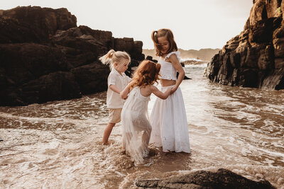 Three children in white clothing stand in the shallow water at a rocky beach, holding hands and smiling as waves lap around them—a perfect moment captured by a talented NSW family photographer.