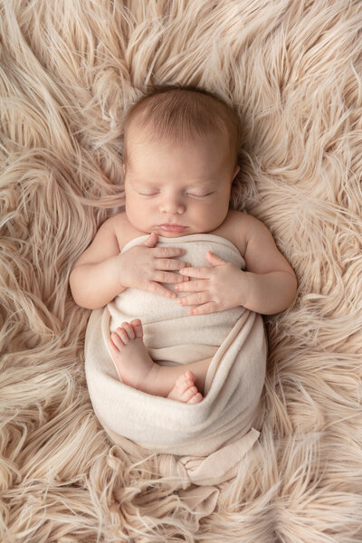timeless newborn portrait of a sleeping newborn with their hands lightly resting on their chest; the baby is lightly swaddled in cream with their folded legs peeking out; the baby is sleeping on a taupe colored mohair rug