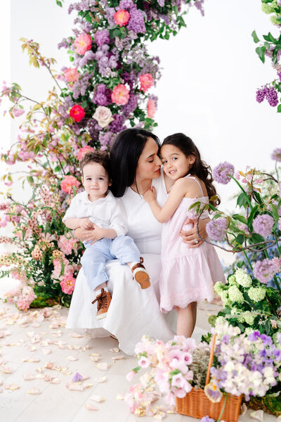 A seated mother hugs her daughter and holds her young son on her lap, all surrounded by abundant pastel blooms and scattered petals on the floor in a cozy, floral-themed studio.