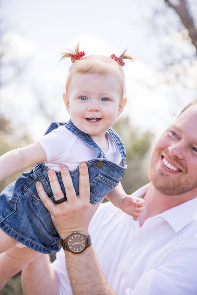 Dad in white shirt holding happy toddler girl with pigtails in the air