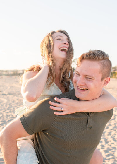 Engagement Photographer, an engaged couple laughs on the beach, she rides on his back