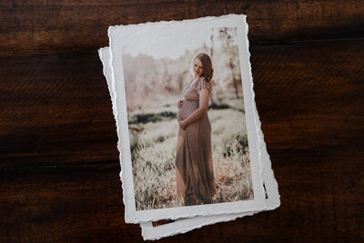A deckled edge print of a maternity photo showing a woman standing in a serene outdoor setting, gently cradling her belly while wearing a light brown dress.