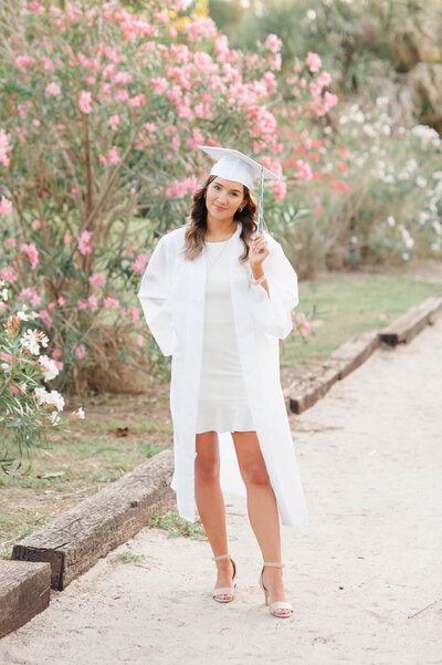 Senior standing on a beach access path in her white cap and gown smiling at the camera while Orlando family photographer M. Lauren takes her photo.