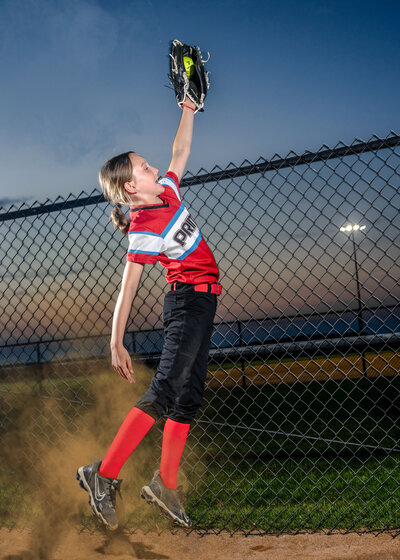 A young girl jumps to catch a softball before it goes over the fence.