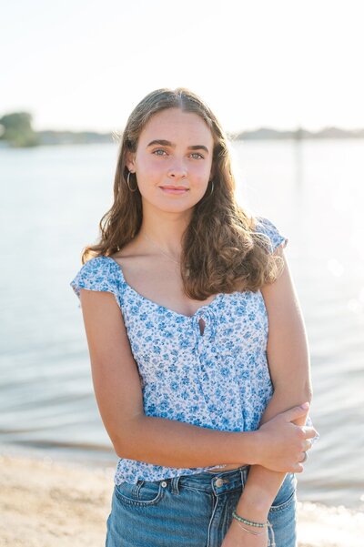 High school senior wearing a blue floral top holds her arms close and softly smiles.