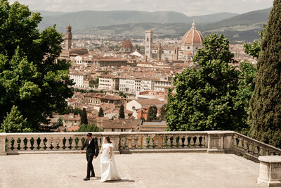 Bride and groom walk hand in hand above Florence with panoramic views of the Duomo, terracotta rooftops, and Tuscan hills — a timeless moment framed by architecture and intimacy.