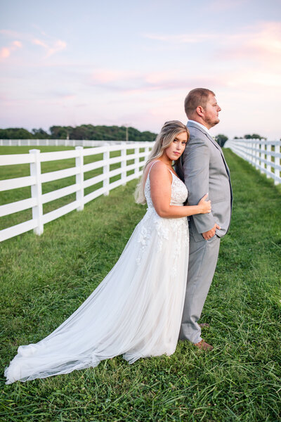 Couple standing in farm field on their wedding day, bride hugging groom from behind while bride looks at camera and groom looks away