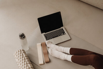 A cozy fitness setup featuring a beige yoga mat, foam roller, and glute bands in neutral tones. A woman’s legs, dressed in deep wine-colored leggings and white socks, rest on the mat. An open laptop and reusable water bottle complete the scene. Captured for Carter Fitness’ social media refresh, this image promotes mindful movement and wellness.