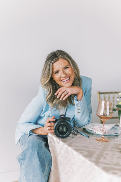 Headshot of Lindsey Kristian wearing a blue blazer and holding her camera with her elbow leaning on a table