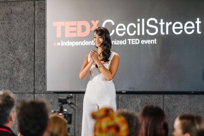 Trang Nova on stage at her TEDx talk wearing a white tailored jumpsuit with her hands held in front of her chest while smiling at the crowd. 