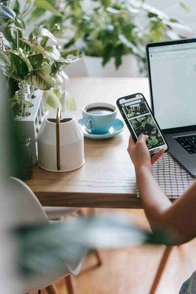 A woman multitasking at home, using a laptop while checking her smartphone, surrounded by a cozy living space.